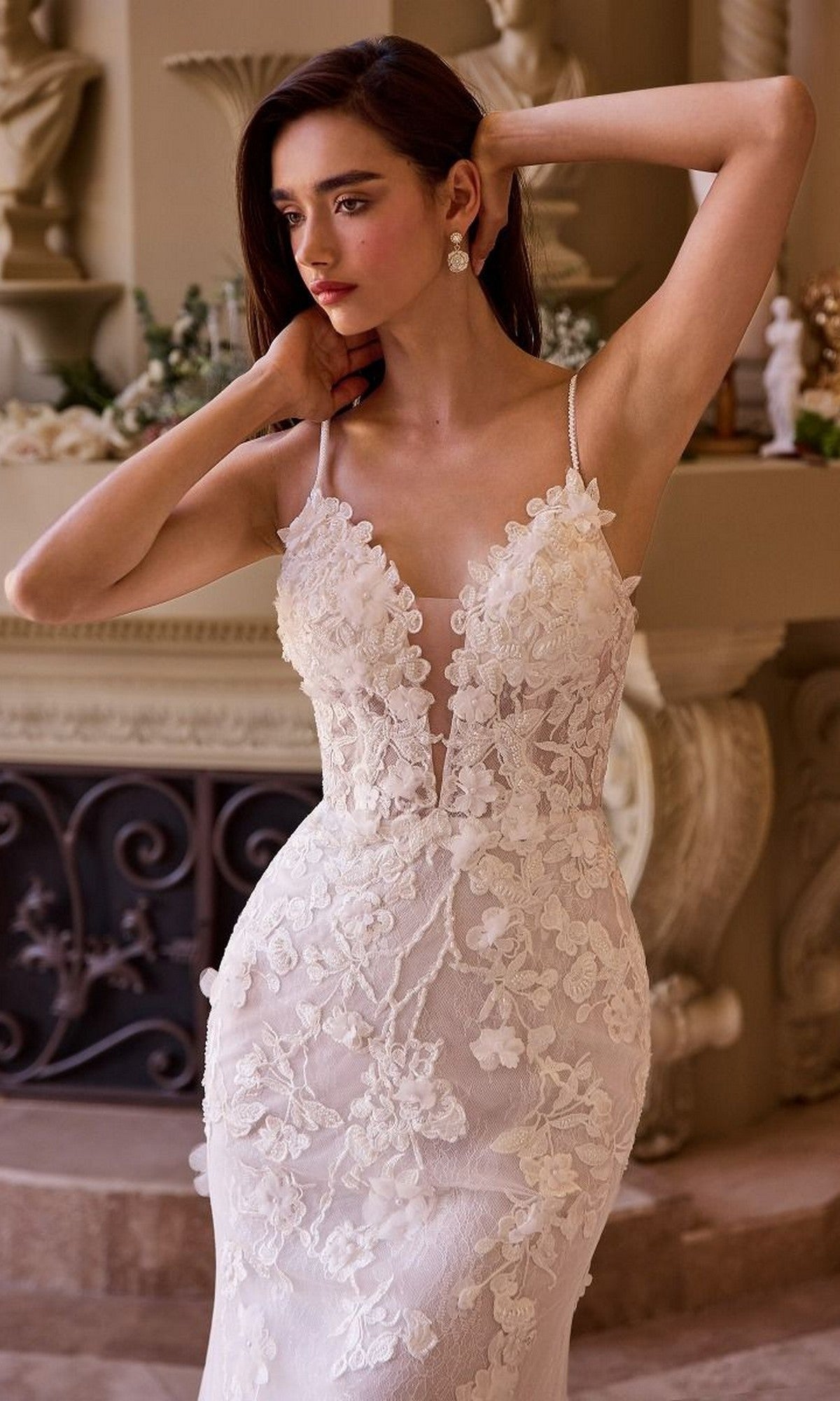 A woman stands indoors before a decorative fireplace, wearing the Andrea and Leo WL053 long wedding dress—a sleeveless lace gown with floral applique, thin straps, and an elegant adjustable lace-up back for a perfect fit.